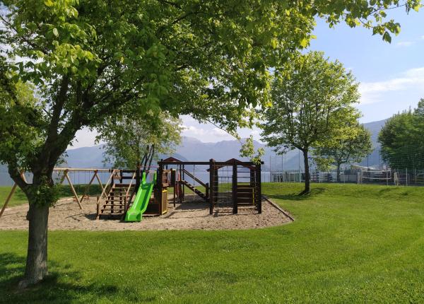 Parco giochi con scivolo verde e altalene, circondato da alberi e montagne.